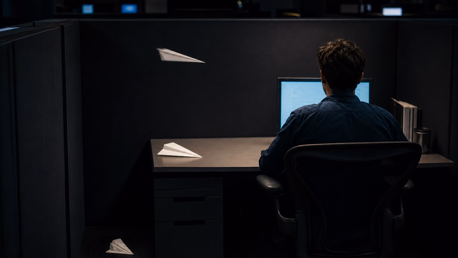 A person works alone in a dark office cubicle while paper airplanes pass unseen around them.